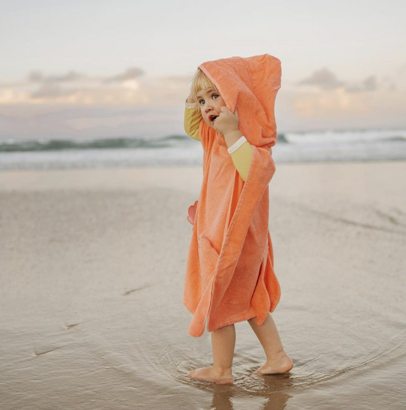 Blonde haired little girl in coral coloured hooded beach towel standing on a beach