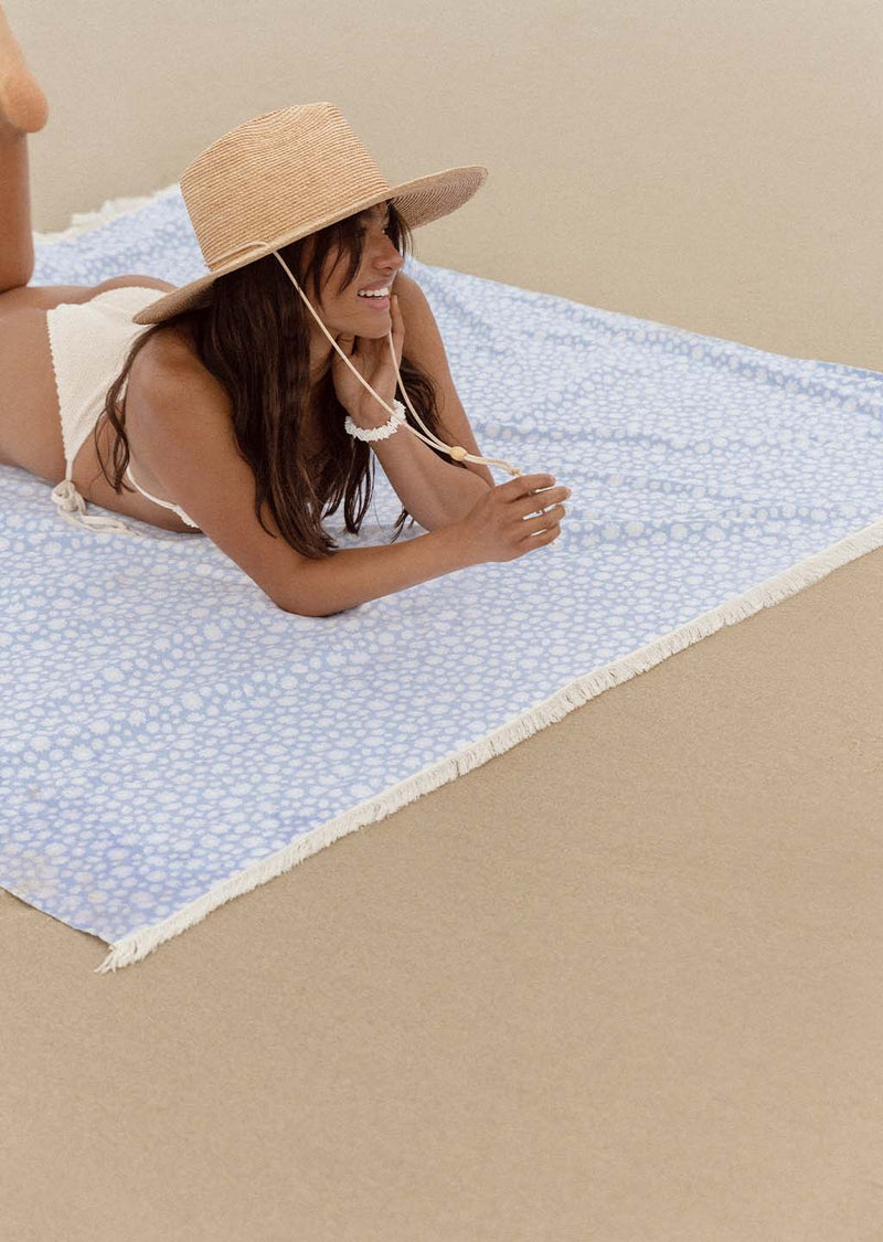 Dark haired lady in straw hat and white bikini lying on blue and white patchy beach towel on top of sand